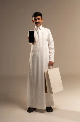 عرض شاشة سوداء فارغة عبر الهاتف المحمول، مفهوم التسوق عن بعد, عمليات الشراء الالكتروني عبر الانترنت, بورتريه لشاب عربي خليجي سعودي يرتدي الزي التقليدي يمسك بيده أكياس ورقية ملونة, التبضع والاستفادة من العروض والتخفيضات, الجسم بالطول الكامل, خلفية رمادية