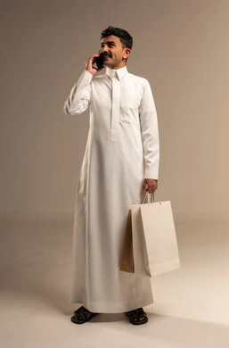 Completing the ordering process using a mobile phone, the concept of remote shopping, online electronic purchases, a portrait of a young Arab Gulf Saudi man wearing traditional attire holding colorful paper bags, shopping and taking advantage of offers and discounts, full-body, gray background.