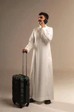 Communicating with family and friends, making phone calls via mobile, a portrait of a smiling young Arab Saudi Gulf man wearing traditional attire holding a mobile phone and a travel bag, direct booking through tourism applications and programs, full-body image, gray background.