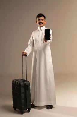 عرض شاشة سوداء فارغة، حجز مباشر عبر تطبيقات وبرامج السياحة, التواصل مع الأهل والأصدقاء, تصفح مواقع التواصل الاجتماعي عبر الجوال, بورتريه لشاب عربي سعودي خليجي مبتسم يرتدي الثوب التقليدي يمسك بيده الهاتف المحمول وحقيبة السفر, الجسم بالطول الكامل, خلفية رمادية