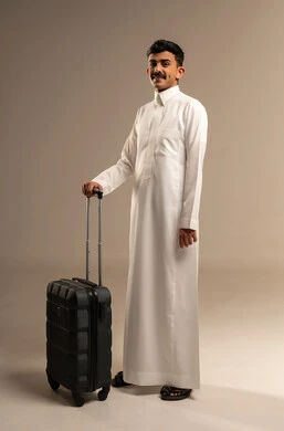 The concept of tourism and taking recreational trips during vacation time, standing upright and waiting for the flight time, a portrait of a smiling Arab Gulf Saudi man wearing traditional attire holding a travel bag in his hand, preparing to start the trip during the summer vacation with joy, full-body, gray background.