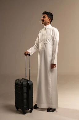 مفهوم السياحة والقيام بالجولات الترفيهية في وقت الإجازة, الوقوف باستقامة وانتظار موعد الطائرة, بورتريه لرجل عربي خليجي سعودي مبتسم يرتدي الثوب التقليدي يمسك بيده حقيبة السفر, الاستعداد لبدء الرحلة في العطلة الصيفيه بسرور, الجسم بالطول الكامل, خلفية رمادية