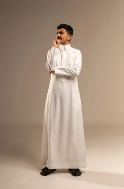 التأمل و التخطيط للمستقبل، التمعن والتركيز لإيجاد حل لمشكلة، بورتريه لشاب عربي خليجي سعودي يرتدي الزي التقليدي  يضع يده على ذقنه بإيماءات التفكير بشيء ما, خلفية رمادية