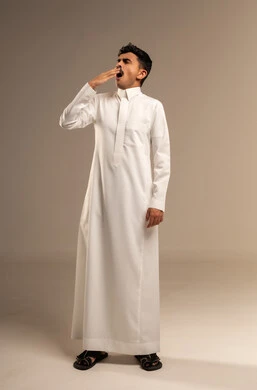 The feeling of tiredness and the need for sleep and rest, the sensation of laziness, lethargy, and exhaustion, a portrait of a young Arab Gulf Saudi man wearing traditional attire, placing his hand over his mouth with gestures of yawning and drowsiness, full-body image, gray background.