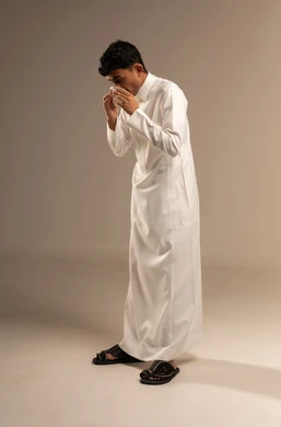 The need for treatment and medication, nasal inflammation and allergic sinuses, feeling sick and having a cold, a portrait of a young Arab Gulf Saudi man wearing traditional attire holding a tissue to his face with sneezing gestures, full body, gray background.