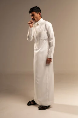 Feeling sick and coughing, infection with the coronavirus, influenza, and pneumonia, a portrait of a young Arab Gulf Saudi teenager wearing traditional attire raising his hand to his mouth in coughing gestures, gray background.