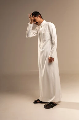 إيماءات تذكر أمر ما بعد فوات الاوان، الشعور بألم في الرأس والجبين، بورتريه لرجل عربي خليجي سعودي يرتدي الزي التقليدي  يضع يده على رأسه وينظر إلى الاسفل بتعابير التفكير، التركيز والتمعن لإيجاد حل لمشكلة معينة، الجسم بالطول الكامل، خلفية رمادية.