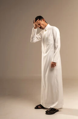 إيماءات تذكر أمر ما بعد فوات الاوان، الشعور بألم في الرأس والجبين، بورتريه لرجل عربي خليجي سعودي يرتدي الزي التقليدي  يضع يده على رأسه وينظر إلى الاسفل بتعابير التفكير، التركيز والتمعن لإيجاد حل لمشكلة معينة، الجسم بالطول الكامل، خلفية رمادية.