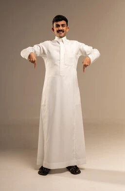 Looking at the camera with gestures of joy, pointing and clarifying something, identifying a specific location, indicating the place of an item, a portrait of a young Arab Gulf Saudi man wearing traditional attire pointing down with his index finger, full-body image, gray background.