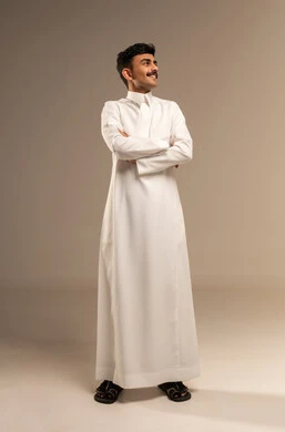 الاناقة والاهتمام بالمظهر الخارجي، الوقوف باستقامة والنظر الى شيء ما بايماءات السرور و السعادة ، بورتريه لشاب عربي خليجي سعودي مبتسم يرتدي  الثوب التقليدي يقف بايدي متقاطعة ، الجسم بالطول الكامل، خلفية رمادية
