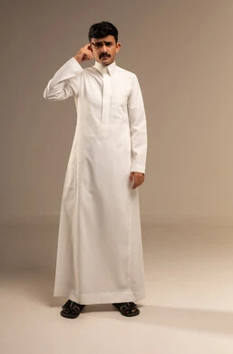 تعابير العبوس والانزعاج، نظرات التحدي والغضب، بورتريه لشاب عربي خليجي سعودي يرتدي الثوب التقليدي ينظر الى الكاميرا يضع يده على وجهه بإيماءات التأمل والتفكير، مفهوم عدم القبول والرضا، الجسم بالطول الكامل، خلفية رمادية