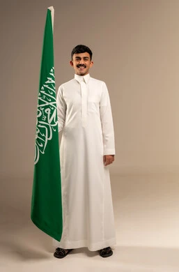 إحياء ذكرى اليوم الوطني السعودي 23 سبتمبر, يوم العلم السعودي 11 مارس، إيماءات الفخر و السرور بالانتماء للوطن, بورتريه لشاب عربي خليجي سعودي مبتسم يرتدي  الثوب التقليدي يحمل بيده علم المملكة، الجسم بالطول الكامل، خلفية رمادية