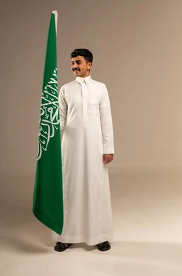 يوم العلم السعودي 11 مارس، إحياء ذكرى اليوم الوطني السعودي 23 سبتمبر, إيماءات الفخر و السرور بالانتماء للوطن, بورتريه لشاب عربي خليجي سعودي مبتسم يرتدي  الثوب التقليدي يحمل بيده علم المملكة، الجسم بالطول الكامل، خلفية رمادية