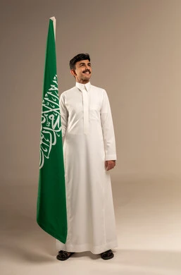 Commemoration of the Saudi National Day on September 23, Saudi Flag Day on March 11, gestures of pride and joy in belonging to the homeland, a portrait of a smiling young Arab Gulf Saudi man wearing traditional attire holding the Kingdom's flag in his hand, full-body, gray background.
