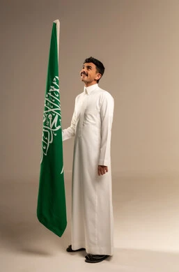 Saudi Flag Day is March 11, commemorating Saudi National Day on September 23, gestures of pride and joy in belonging to the homeland, a portrait of a smiling young Arab Gulf Saudi man wearing traditional attire holding the Kingdom's flag in his hand, full-body, gray background.