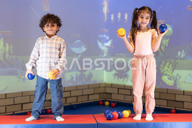 Expressions of joy and happiness, moments of fun and excitement, spending happy times at an amusement park, a cheerful interactive atmosphere, an Emirati Gulf Arab girl enjoying an interactive throwing game with her brother, an amusement park.