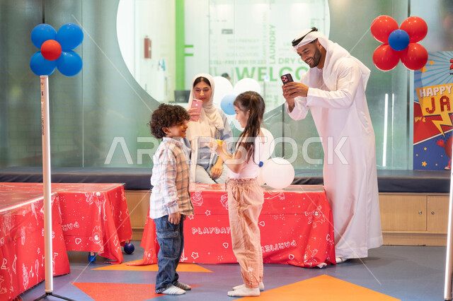 رجل اماراتي يلتقط صور السيلفي ، أجواء تفاعلية مبهجة ،قضاء الاوقات السعيده مع الابناء ، استخدام الجوال لتوثيق اللحظات التذكاريه ،عائلة عربية خليجية إماراتية تستمتع بوقت ممتع في لعبة الرمي التفاعلية،  مركز ألعاب ترفيهي، تعابير السرور والسعادة، مشاركة الأب والأم أطفالهما لحظات المرح والحماس