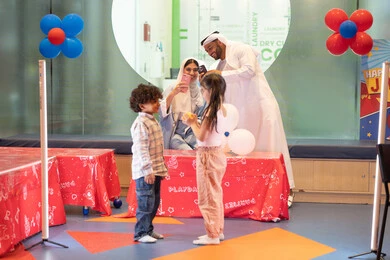 Spending happy times with children, using a mobile phone to document memorable moments, an Emirati Gulf Arab family enjoying a fun time playing the interactive archery game, an Emirati man taking selfies, a joyful interactive atmosphere, an entertainment center, expressions of joy and happiness, and a father and mother sharing moments of fun with their children. Enthusiasm