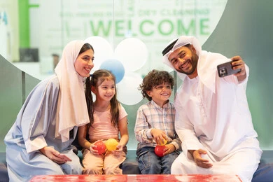 A cheerful interactive atmosphere, spending happy times with the children, an entertainment games center, using the mobile phone to document memorable moments, expressions of pleasure and happiness, an Emirati man taking selfies, the father and mother sharing moments of fun and excitement with their children, an Emirati Gulf Arab family enjoying a fun time in the interactive throwing game.