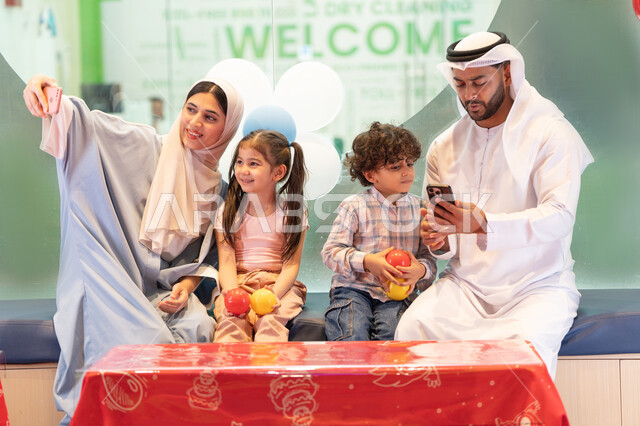 Entertainment games center, using mobile phones to document memorable moments, a cheerful interactive atmosphere, expressions of pleasure and happiness, an Emirati woman taking selfies, father and mother sharing moments of fun and excitement with their children, an Emirati Gulf Arab family enjoying a fun time in the interactive throwing game, spending happy times with the children