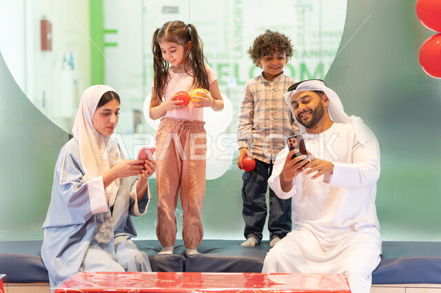 مشاركة الأب والأم أطفالهما لحظات المرح والحماس ،  التقاط صور السيلفي ، مركز ألعاب ترفيهي،استخدام الجوال لتوثيق اللحظات التذكاريه ، أجواء تفاعلية مبهجة ، عائلة عربية خليجية إماراتية تستمتع بوقت ممتع في لعبة الرمي التفاعلية ،تعابير السرور والسعادة،  قضاء الاوقات السعيده مع الابناء