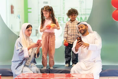 Parents sharing moments of fun and excitement with their children, taking selfies, an entertainment game center, using mobile phones to document memorable moments, a cheerful interactive atmosphere, an Emirati Gulf Arab family enjoying a fun time in the interactive throwing game, expressions of pleasure and happiness, spending happy times with the children