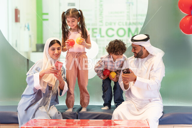 التقاط صور السيلفي ، مركز ألعاب ترفيهي،استخدام الجوال لتوثيق اللحظات التذكاريه ، مشاركة الأب والأم أطفالهما لحظات المرح والحماس ،  أجواء تفاعلية مبهجة ، عائلة عربية خليجية إماراتية تستمتع بوقت ممتع في لعبة الرمي التفاعلية ،تعابير السرور والسعادة،  قضاء الاوقات السعيده مع الابناء