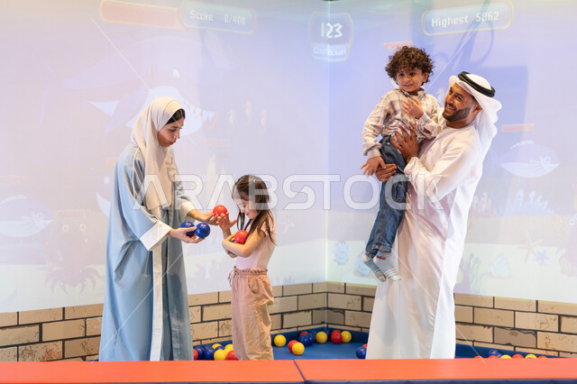 أجواء تفاعلية مبهجة ، اب اماراتي يحمل ابنه لاعلى بتعابير البهجه ، عائلة عربية خليجية إماراتية تستمتع بوقت ممتع في لعبة الرمي التفاعلية ،تعابير السرور والسعادة،  قضاء الاوقات السعيده مع الابناء ،مركز ألعاب ترفيهي، مشاركة الأب والأم أطفالهما لحظات المرح والحماس
