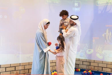 تعابير السرور والسعادة،  قضاء الاوقات السعيده مع الابناء ،مركز ألعاب ترفيهي، مشاركة الأب والأم أطفالهما لحظات المرح والحماس ، أجواء تفاعلية مبهجة ،عائلة عربية خليجية إماراتية تستمتع بوقت ممتع في لعبة الرمي التفاعلية