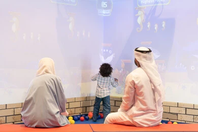 قضاء الاوقات السعيده مع الابناء ،مركز ألعاب ترفيهي، مشاركة الأب والأم أطفالهما لحظات المرح والحماس ، أجواء تفاعلية مبهجة ،تعابير السرور والسعادة،  صوره من الخلف لعائلة عربية خليجية إماراتية تستمتع بوقت ممتع في لعبة الرمي التفاعلية