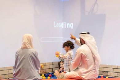 مركز ألعاب ترفيهي، مشاركة الأب والأم أطفالهما لحظات المرح والحماس ، أجواء تفاعلية مبهجة ،تعابير السرور والسعادة،  صوره من الخلف لعائلة عربية خليجية إماراتية تستمتع بوقت ممتع في لعبة الرمي التفاعلية ، قضاء الاوقات السعيده مع الابناء