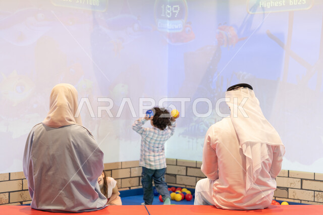 أجواء تفاعلية مبهجة ،تعابير السرور والسعادة، مشاركة الأب والأم أطفالهما لحظات المرح والحماس ،  مركز ألعاب ترفيهي، صوره من الخلف لعائلة عربية خليجية إماراتية تستمتع بوقت ممتع في لعبة الرمي التفاعلية ، قضاء الاوقات السعيده مع الابناء