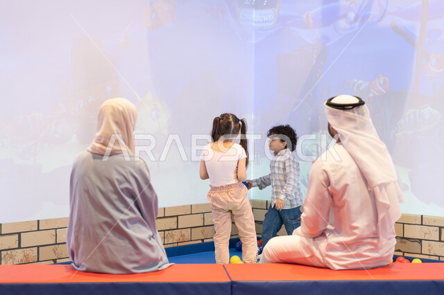 تعابير السرور والسعادة، مشاركة الأب والأم أطفالهما لحظات المرح والحماس ،  مركز ألعاب ترفيهي، صوره من الخلف لعائلة عربية خليجية إماراتية تستمتع بوقت ممتع في لعبة الرمي التفاعلية ، أجواء تفاعلية مبهجة ،قضاء الاوقات السعيده مع الابناء
