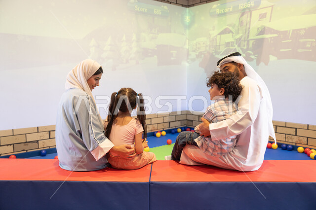 صوره من الخلف لعائلة عربية خليجية إماراتية تستمتع بوقت ممتع في لعبة الرمي التفاعلية ، أجواء تفاعلية مبهجة ،قضاء الاوقات السعيده مع الابناء ، مركز ألعاب ترفيهي، مشاركة الأب والأم أطفالهما لحظات المرح والحماس ، تعابير السرور والسعادة
