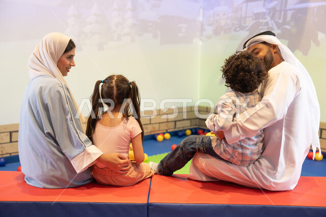 A cheerful interactive atmosphere, spending happy times with the children, a photo from the back of an Emirati Gulf Arab family enjoying a fun time in the interactive throwing game, an entertainment games center, the father and mother sharing moments of fun and excitement with their children, expressions of pleasure and happiness