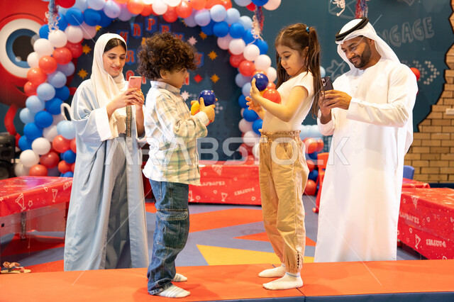 مفهوم التسلية والترفيه، دمج التقنيات المتطوره فى الحياه اليوميه، مشاعر المتعة والفرح، قضاء أوقات سعيده في أرض المرح، المراكز الترفيهيه المسلية في الامارات العربية،عائله عربيه خليجيه اماراتيه تستمتع باللعب فى مدينة الملاهي ، تلعب بالكور الملونه ، استخدام الجوال فى توثيق اللحظات التذكاريه