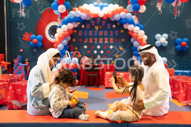 Documenting memorable moments, entertaining entertainment centers in the UAE, spending happy times in Funland, an Emirati Gulf Arab girl enjoying playing at an amusement park with her family and using a mobile phone, feelings of joy and happiness, the concept of entertainment and recreation, integrating advanced technologies into daily life.