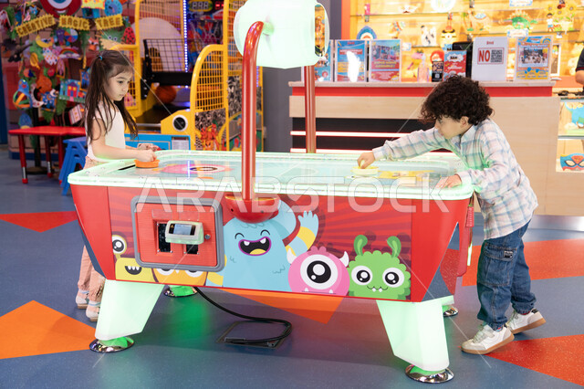 طاولة مضيئة بألوان زاهية ،ايماءات السرور والسعاده ،أجواء مليئة بالمرح والحماس، مراكز الالعاب الترفيهيه في الامارات،  قضاء اوقات ممتعه ،  ولد عربي خليجي إماراتي يرتدي الزى الكاجوال ، يشارك اخته بلعب الهوكي الهوائي فى مدينه الالعاب