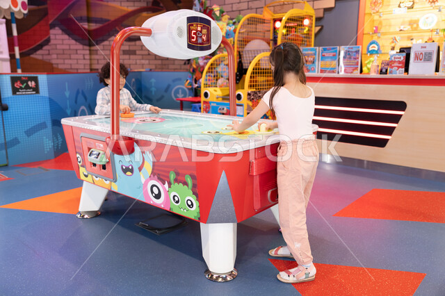 ايماءات السرور والسعاده ،أجواء مليئة بالمرح والحماس، مراكز الالعاب الترفيهيه في الامارات،  طاولة مضيئة بألوان زاهية ، قضاء اوقات ممتعه ،  ولد عربي خليجي إماراتي يرتدي الزى الكاجوال ، يشارك اخته بلعب الهوكي الهوائي فى مدينه الالعاب