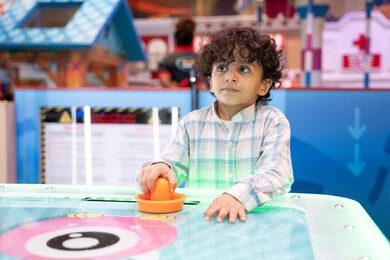 أجواء مليئة بالمرح والحماس، مراكز الالعاب الترفيهيه في الامارات، طاولة مضيئة بألوان زاهية ، قضاء اوقات ممتعه ، ولد عربي خليجي إماراتي يرتدي الزى الكاجوال ، يستمتع بلعب الهوكي الهوائي فى مدينه الالعاب