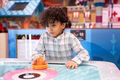 مراكز الالعاب الترفيهيه في الامارات، طاولة مضيئة بألوان زاهية ، قضاء اوقات ممتعه ، أجواء مليئة بالمرح والحماس، ولد عربي خليجي إماراتي يرتدي الزى الكاجوال ، يستمتع بلعب الهوكي الهوائي فى مدينه الالعاب