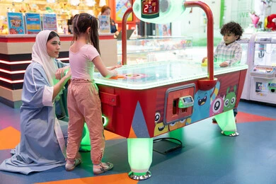 Fun sports activities, challenging and exciting competition, entertainment centers in the UAE, spending happy times with family members, a colorfully illuminated table, an atmosphere full of fun and joy, an Emirati Gulf Arab family enjoying air hockey in an amusement park.