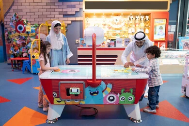 الاستماع بالاوقات السعيده مع افراد الاسره ،ايماءات التحدي والإثارة ،  أجواء مليئة بالمرح والسرور، الأنشطة الرياضية الممتعة، مراكز الالعاب الترفيهيه في الامارات، طاولة مضيئة بألوان زاهية ، عائله عربية خليجية إماراتية  تستمتع بلعب الهوكي الهوائي فى مدينه الالعاب