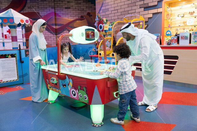 A challenging and exciting competition, fun sports activities, a colorful illuminated table, entertainment centers in the UAE, spending happy times with family members, an atmosphere full of fun and joy, an Arab Gulf Emirati family enjoying playing air hockey in an amusement park.