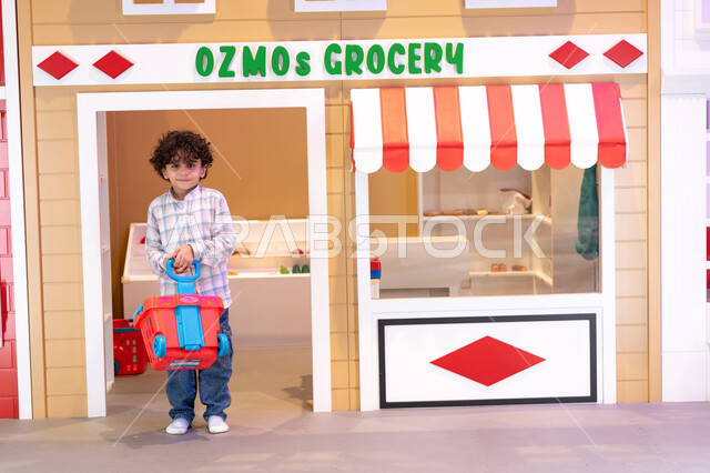 النظر الى الكاميرا بايماءات السعاده، اللعب في قسم البقالة، قضاء وقت ممتع في مركز الالعاب الترفيهيه،  تعلم تسوق الخضار الطازجة ، دعم وتنمية مهارات الأطفال،ولد عربي خليجي اماراتي يرتدي الزى الكاجوال، يجر عربه التسوق، طعام صحي ومفيد