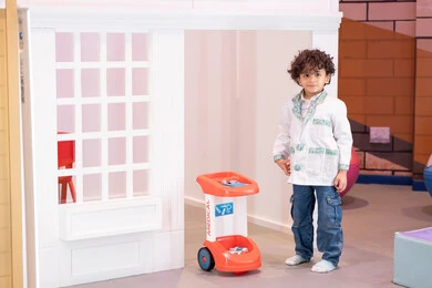 A fun experience simulating the work of doctors, playing professional roles, the concept of education through play, an Arab Gulf Emirati boy wearing a medical coat, playing with a medical toy cart in a children's entertainment center, gestures of innocence and fun, promoting the values ​​of responsibility and caring for others, developing mental and imaginative skills in children