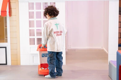 مفهوم التعليم عن طريق اللعب ، تقمص الأدوار المهنية، تجربة ممتعة تحاكي عمل الأطباء، صورة من الخلف  لولد عربي خليجي اماراتي يرتدي معطف طبي ، يلعب بعربة ألعاب طبية في أحد المراكز الترفيهية للأطفال، ايماءات البراءة والمرح، تعزيز قيم المسؤولية والاهتمام بالآخرين ، تنمية المهارات الذهنية والخيالية لدى الأطفال