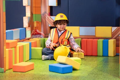 An Emirati Gulf Arab boy wearing a helmet and a safety vest assembling blocks, enhancing mental and intellectual skills, entertainment centers in the Emirates, spending fun times playing and concentrating, caring for and developing children's skills