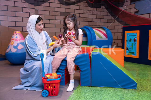 ايماءات المرح والسرور،  الاهتمام ودعم الابناء،  تعزيز مفهوم الغذاء الصحي والمتوازن، تنمية مهارات الأطفال، سيدة عربية خليجية إماراتية ترتدي العباءة والحجاب تشارك ابنتها لحظات ممتعه في مركز الألعاب الترفيهية، قضاء وقت عائلي ممتع مليء بالحب ، تعلم تسوق المنتجات الطازجة من خلال سلة مليئة بالخضار والفواكه البلاستيكية