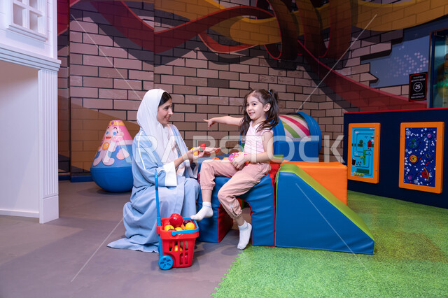 Spending fun family time full of love, learning how to shop for fresh produce through a basket full of plastic fruits and vegetables, promoting the concept of healthy and balanced nutrition, supporting and developing children's skills, an atmosphere full of fun and happiness, an Emirati Gulf Arab woman wearing an abaya and hijab sharing fun moments with her daughter in the entertainment games center